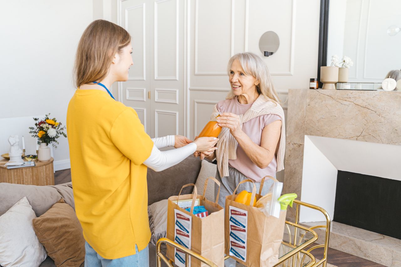 A caregiver bringing groceries to a smiling senior woman in her bright living room