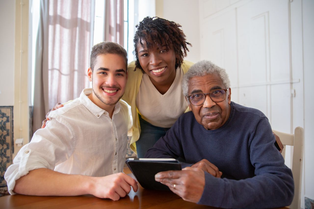 A caregiver smiling warmly beside a senior man and a younger family member gathered around a tablet at home